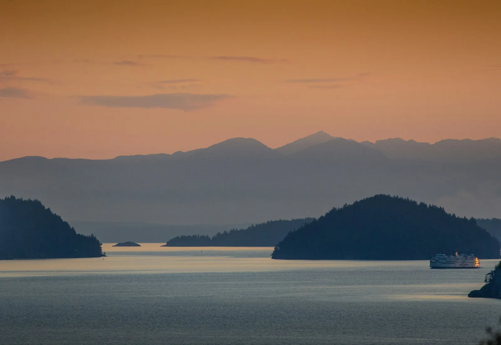 Dark wooded islands in layers on a glassy bay at dusk, a ferry far off and orange sky above